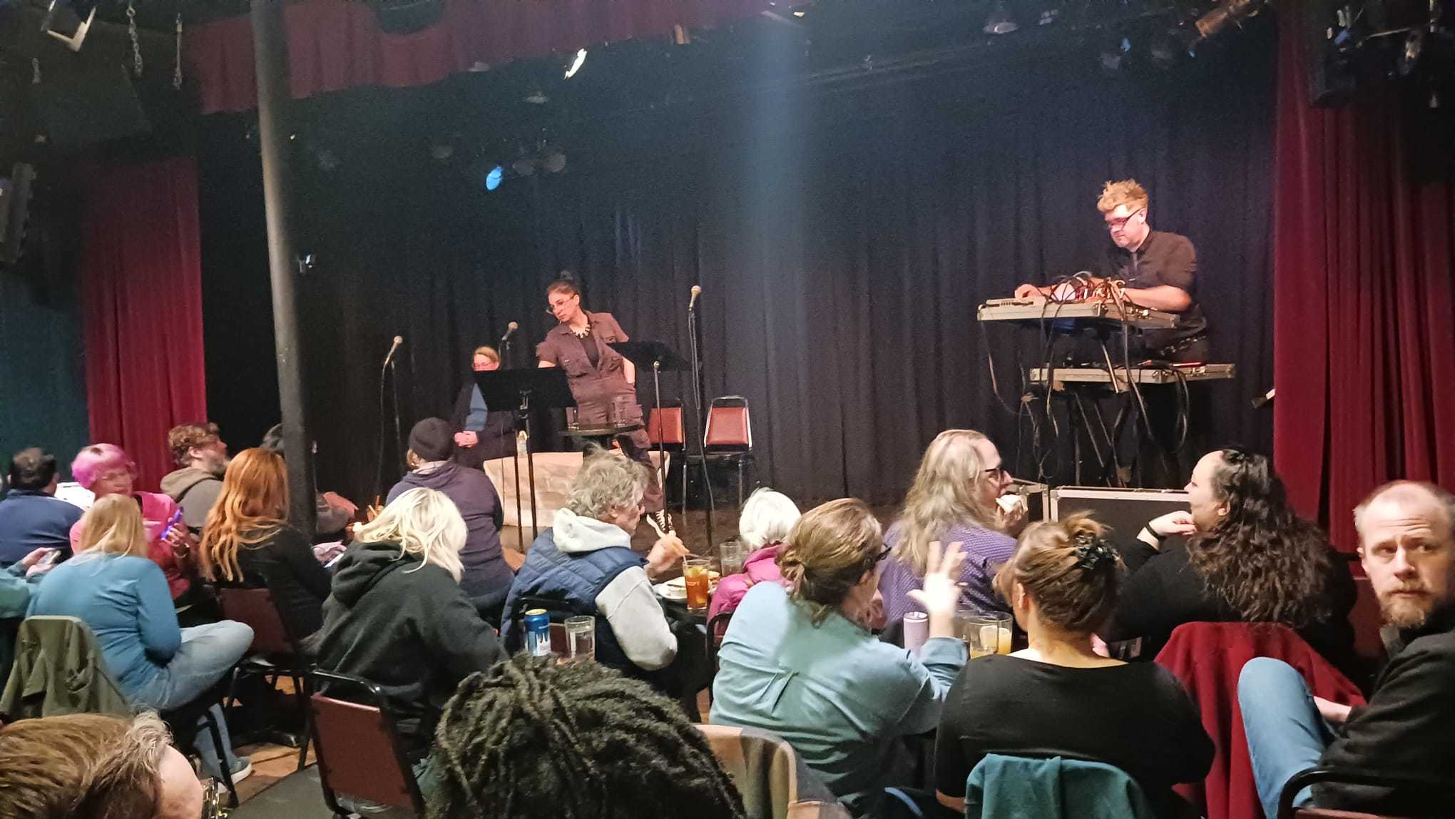 A crowd of people at Bryant Lake Bowl waiting for the show to start. Naomi Kritzer is seated in the far left--mostly obscured by a podium/table.